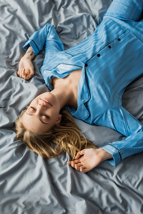 top view of carefree woman in blue silk pajama lying with closed eyes on bed in modern apartment