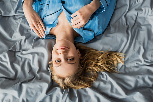 top view of happy woman in blue silk pajama lying on bed and looking at camera