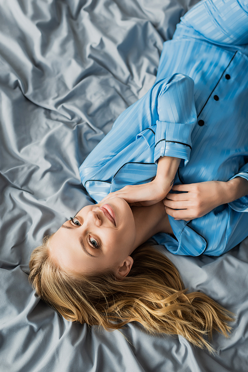 top view of cheerful woman in blue silk pajama lying on bed and looking at camera