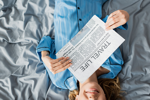 top view of cropped woman smiling while reading travel life newspaper in bed