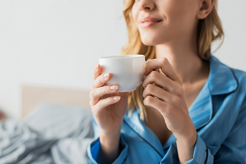 cropped view of pleased young woman in blue pajama holding cup of coffee