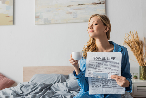 pleased young woman holding travel life newspaper and cup of coffee in bedroom