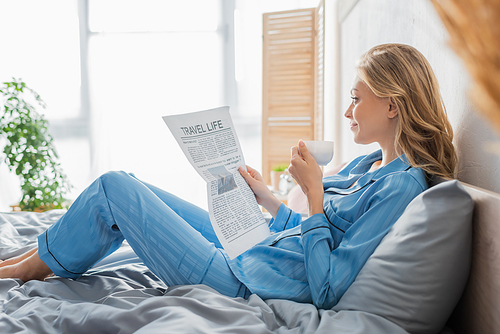 side view of carefree young woman reading travel life newspaper and holding cup of coffee in bed