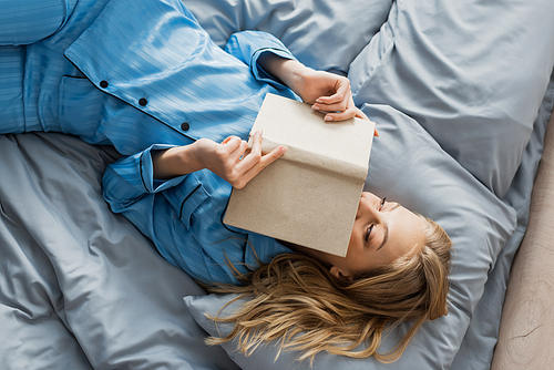 top view of satisfied young woman in blue silk nightwear covering face with book in bed