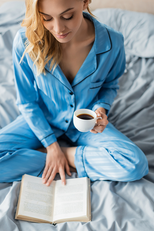 overhead view of young woman in blue silk nightwear reading book while holding cup of coffee