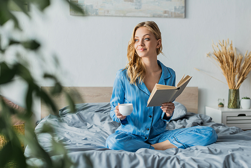 pretty young woman in blue silk nightwear holding book and cup of coffee in modern bedroom