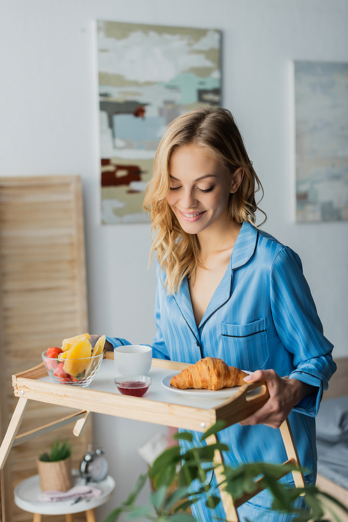 happy woman in blue pajama looking at tray with breakfast in bedroom