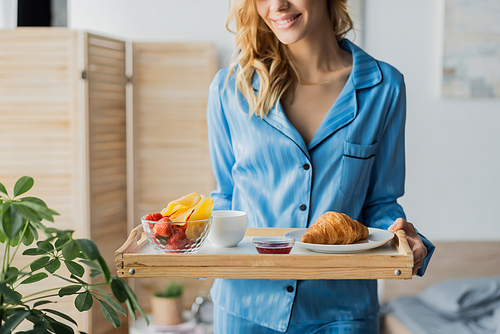 cropped view of happy woman in blue nightwear holding tray with breakfast in bedroom