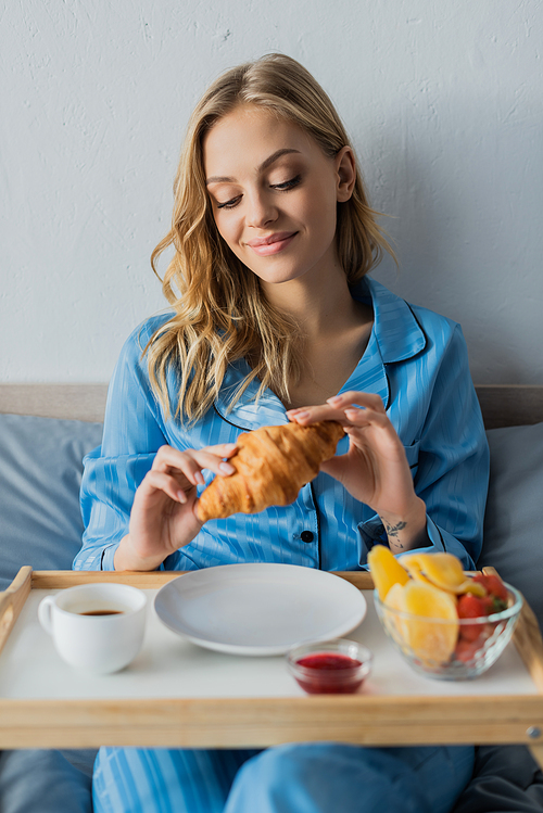 smiling young woman in blue pajama holding fresh croissant near tray while having breakfast in bed