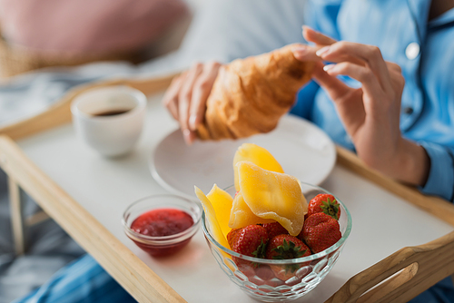 cropped view of woman holding fresh croissant near tray with jam and dried mango while having breakfast in bed