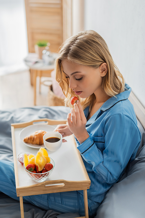 blonde young woman in pajama holding cup of coffee and fresh strawberry near tray while having breakfast in bed