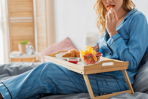 cropped view of woman holding cup of coffee and eating fresh strawberry near tray while having breakfast in bed