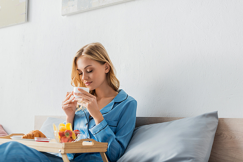 pretty woman holding cup while enjoying coffee flavor near breakfast tray in bed