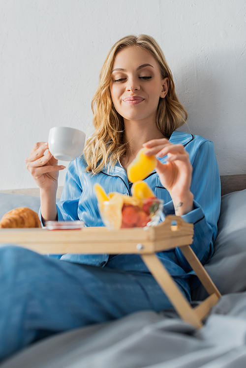 smiling young woman in pajama holding cup of coffee and dried mango near tray while having breakfast in bed