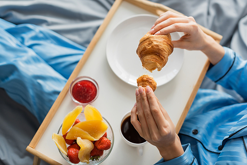 top view of cropped woman holding fresh croissant near tray while having breakfast in bed