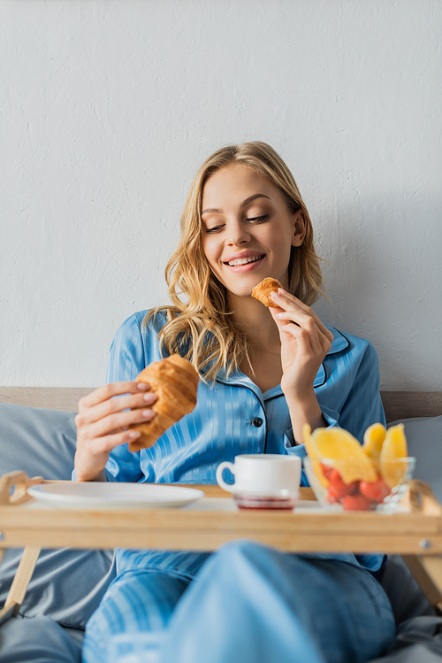 happy woman in pajama eating fresh croissant near tray while having breakfast in bed