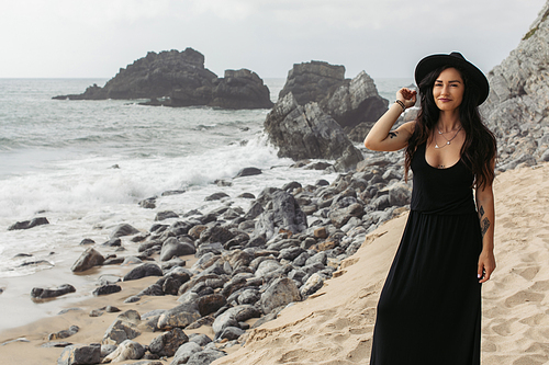 cheerful and tattooed woman in black dress and hat standing on sandy beach in portugal