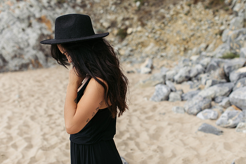 tattooed woman in dress and black hat standing on sandy beach in portugal