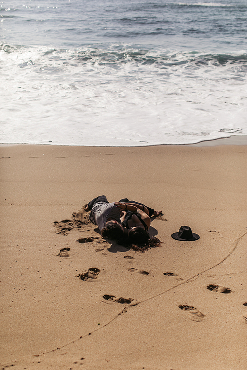 couple hugging and lying on wet sand near ocean on beach