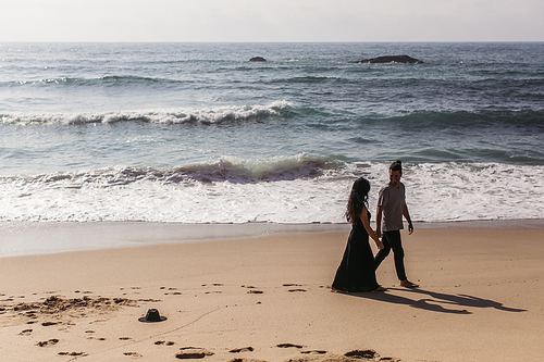 man and woman in dress holding hands while walking on wet sand on beach near ocean