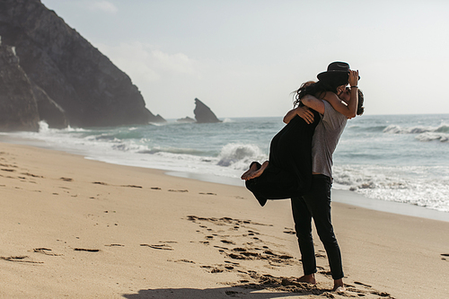 bearded man lifting happy woman in hat and dress on beach near ocean
