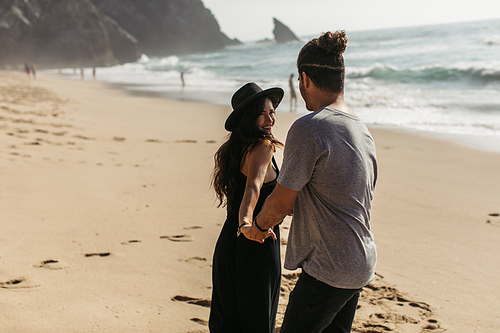 happy woman in hat and black dress holding hand of boyfriend on beach in portugal