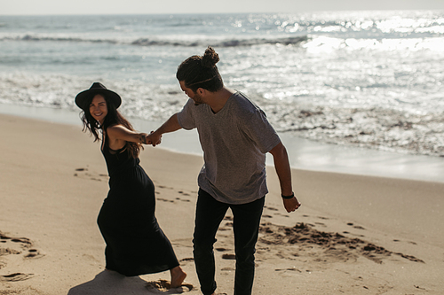cheerful woman in hat and black dress pulling hand of boyfriend on beach in portugal