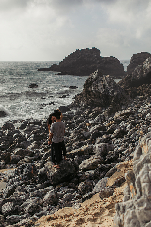 woman wearing hat on boyfriend while standing on rocks near ocean
