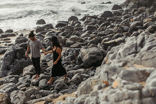 bearded man smiling and holding hands with girlfriend in dress and hat while walking on rocks near ocean