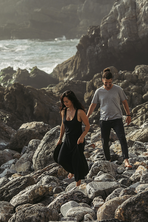bearded man smiling while walking on rocks near tattooed girlfriend in dress near ocean