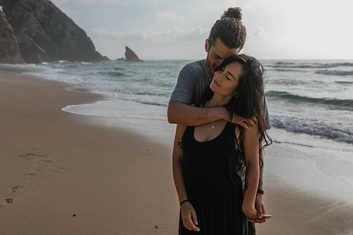 happy man hugging tattooed girlfriend in dress near ocean in portugal