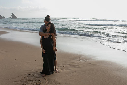 full length of happy man hugging tattooed girlfriend in dress near ocean in portugal