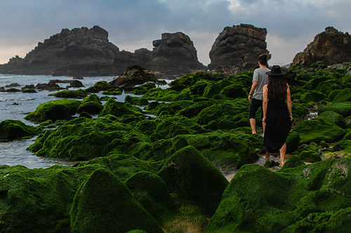 back view of tattooed woman in hat and dress standing behind man on mossy stones near ocean