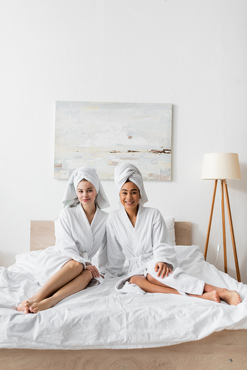 full length of young barefoot women in white soft robes and towels sitting on bed and smiling at camera