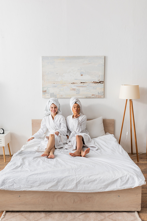 full length of african american woman blowing air kiss near cheerful friend in bathrobe and towel on bed at home