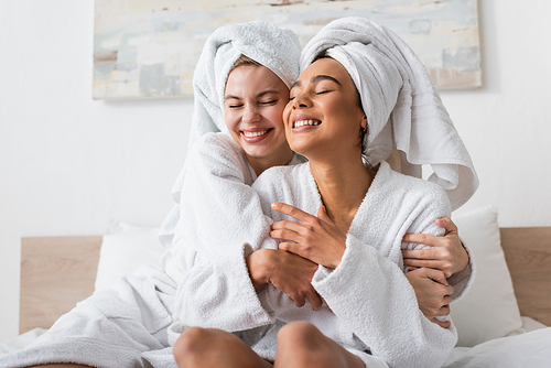 joyful interracial women in white terry bathrobes and towels embracing with closed eyes in bedroom