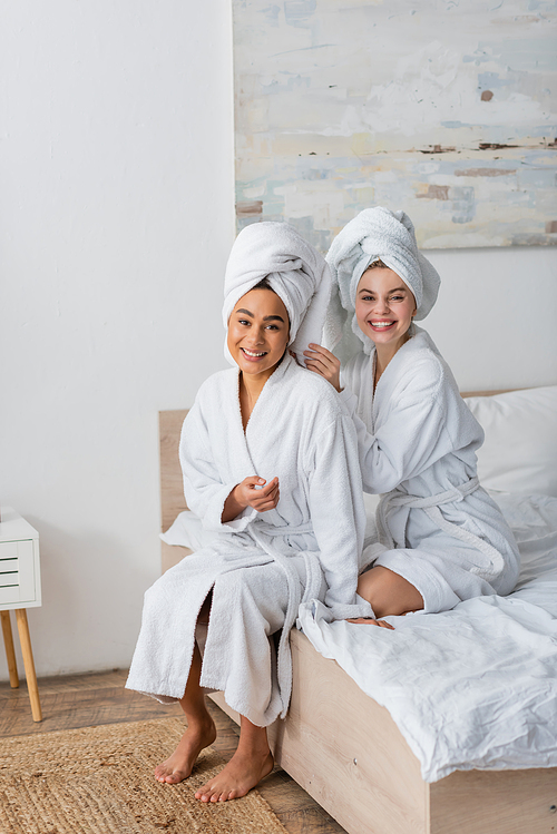 happy interracial women in white soft robes and towels looking at camera while sitting on bed