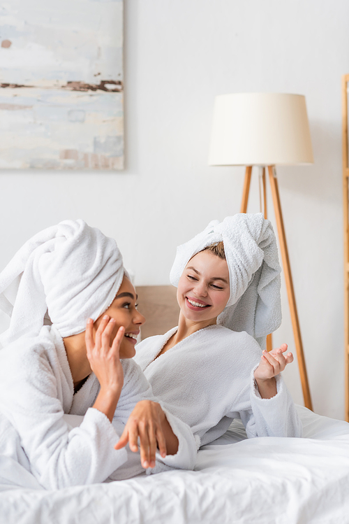 joyful interracial women in white bathrobes and towels gesturing during conversation in bedroom