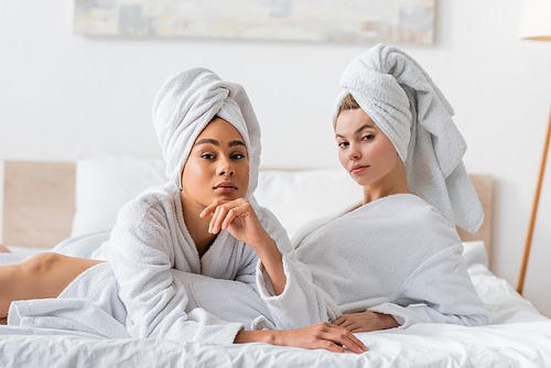 young and pretty interracial women in white terry bathrobes and towels looking at camera on bed at home