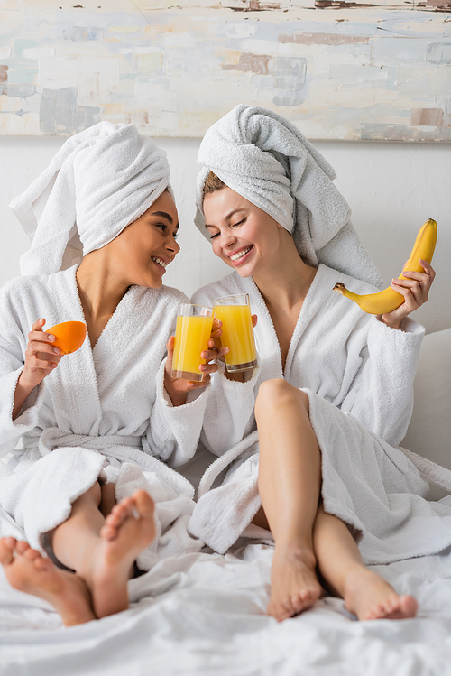 full length of barefoot interracial women in white robes clinking glasses of orange juice while sitting on bed