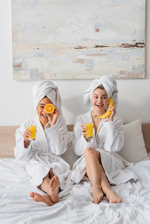 full length of cheerful interracial women with fruits and orange juice having fun while sitting on bed