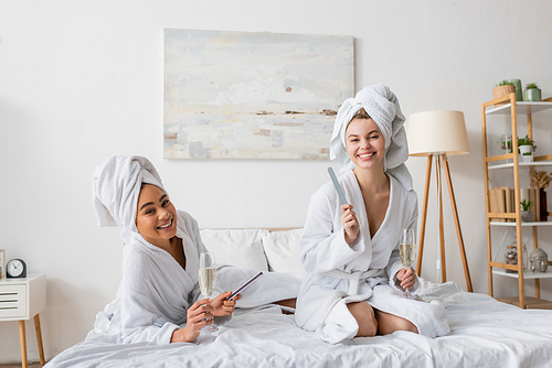 cheerful interracial women with champagne glasses and nail files looking at camera on bed