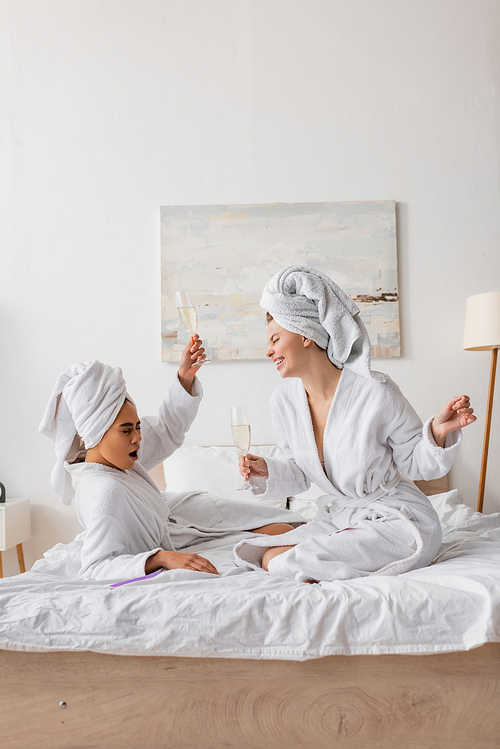 excited african american woman toasting with champagne near happy friend in white robe and towel in bedroom