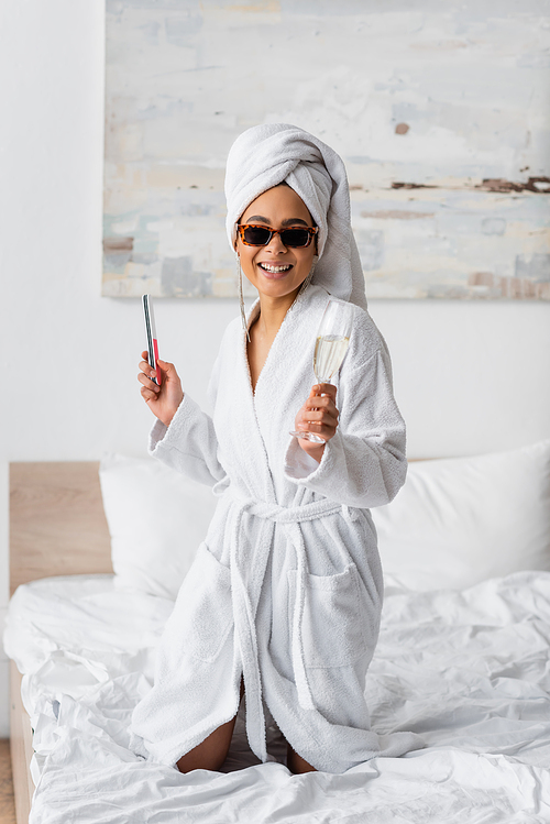 cheerful african american woman in white bathrobe and sunglasses posing with nail file and champagne glass in bedroom