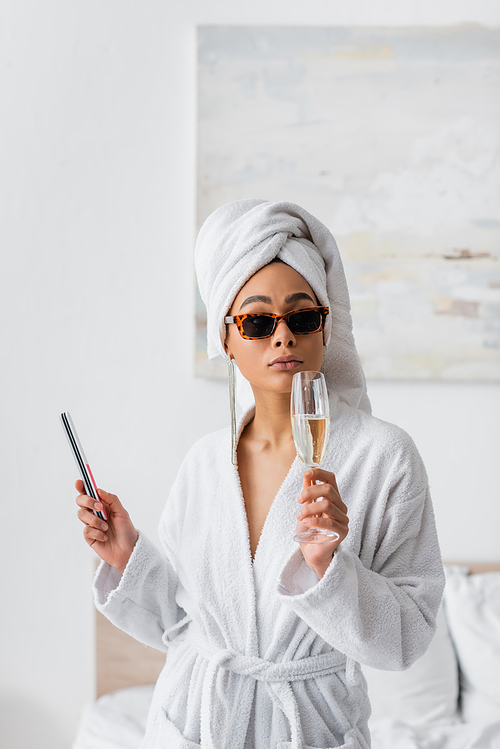 young african american woman in white bathrobe and stylish sunglasses holding champagne glass and nail file in bedroom