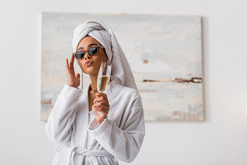 african american woman in terry bathrobe and towel adjusting trendy sunglasses while pouting lips and holding champagne in bedroom