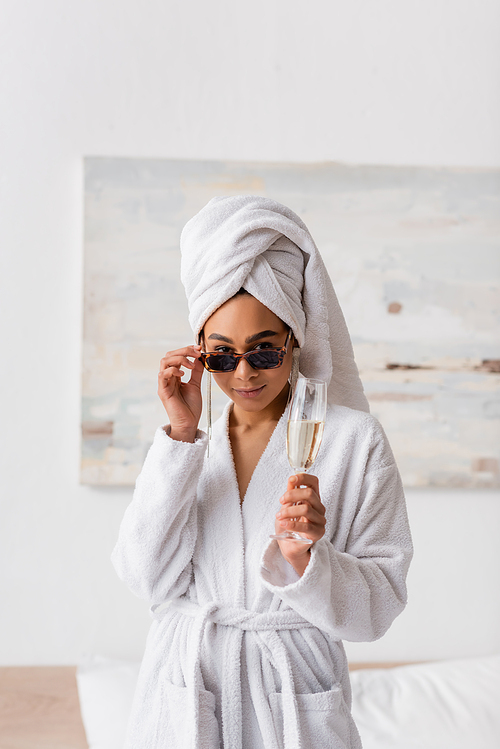 young african american woman in white bathrobe and towel holding champagne and looking at camera over sunglasses in bedroom