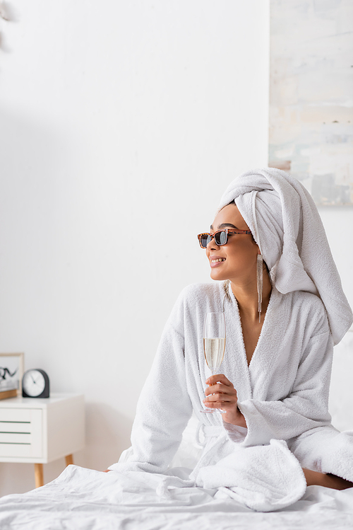 smiling african american woman in terry bathrobe and trendy sunglasses holding champagne and looking away in bedroom