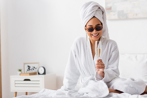 happy african american woman in terry bathrobe and trendy sunglasses sitting on bed with glass of champagne