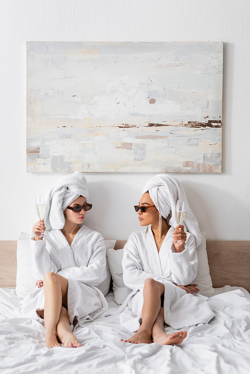 full length of barefoot interracial women in bathrobes and trendy sunglasses holding champagne and looking at each other on bed